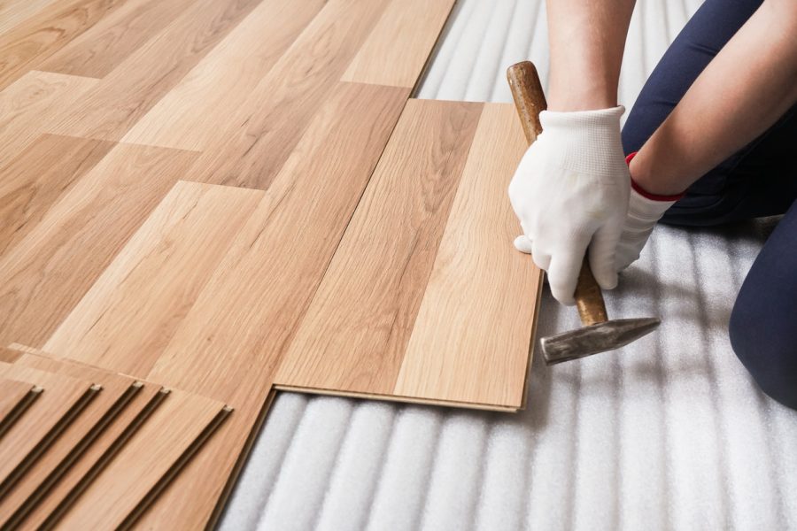 Installing laminated floor, detail on man hands fixing one tile with hammer, over white foam base layer.